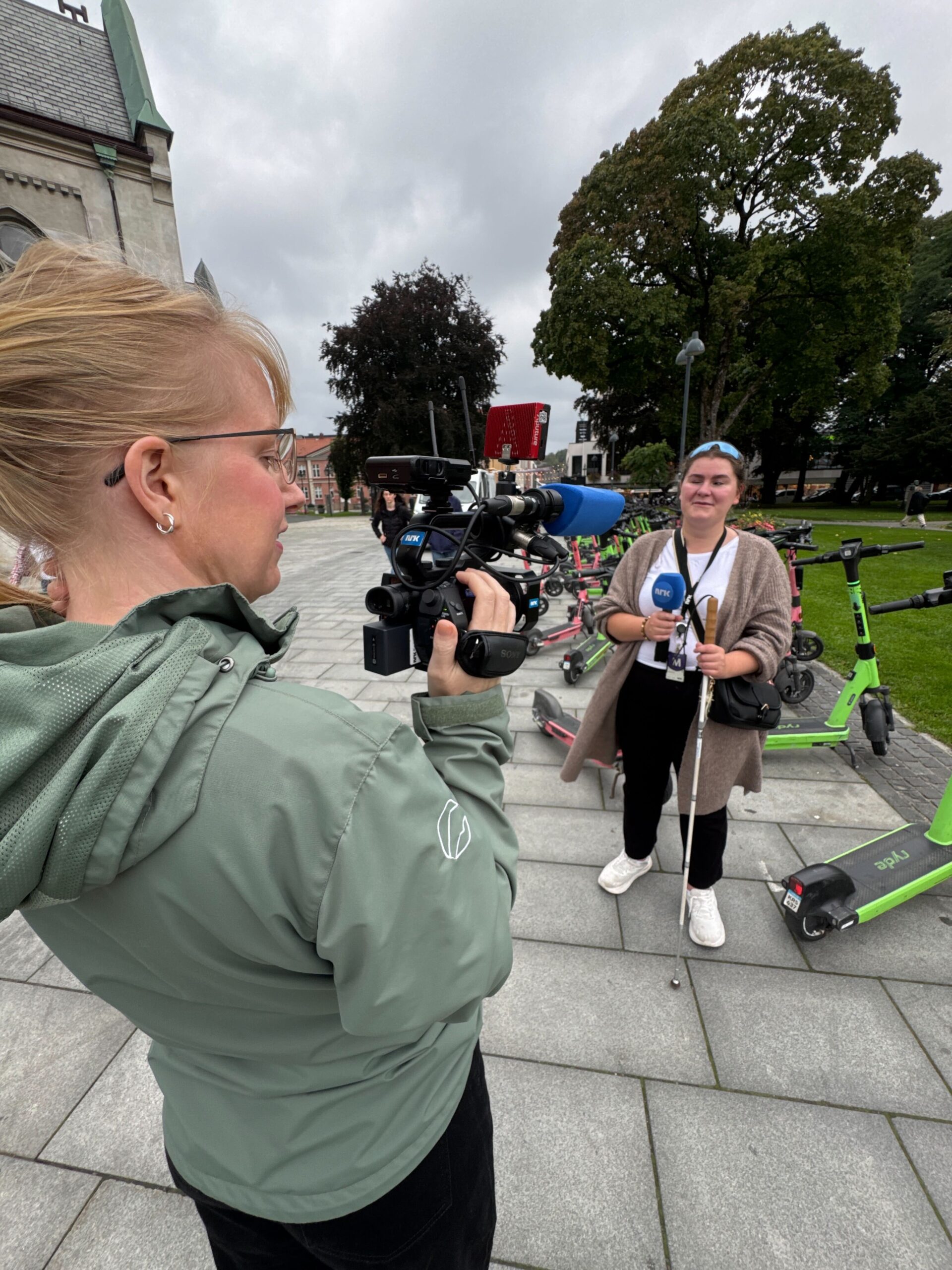 På bildet ser vi Marit stå med ryggen til. Hun holder et lite kamera med NRK-logo på. Kameraet er rette mot Ragna som står noen meter unna med NRK-mikrofon i hånden. 