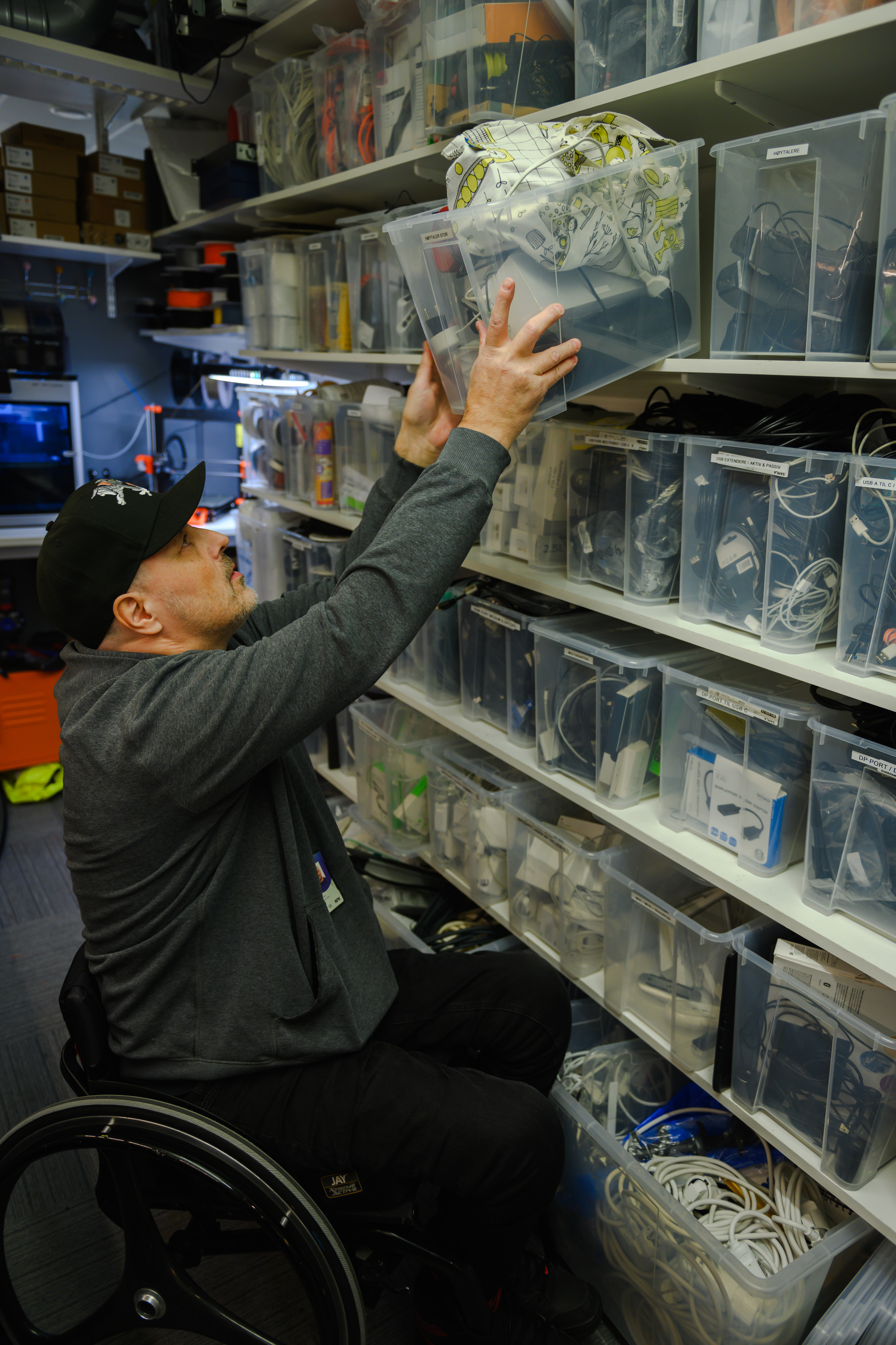 En mann i rullestol henter ned en plasteske med utstyr fra en hylle på en vegg dekket av mange plastbokser fylt med ulikt teknisk utstyr og ledninger og kabler..