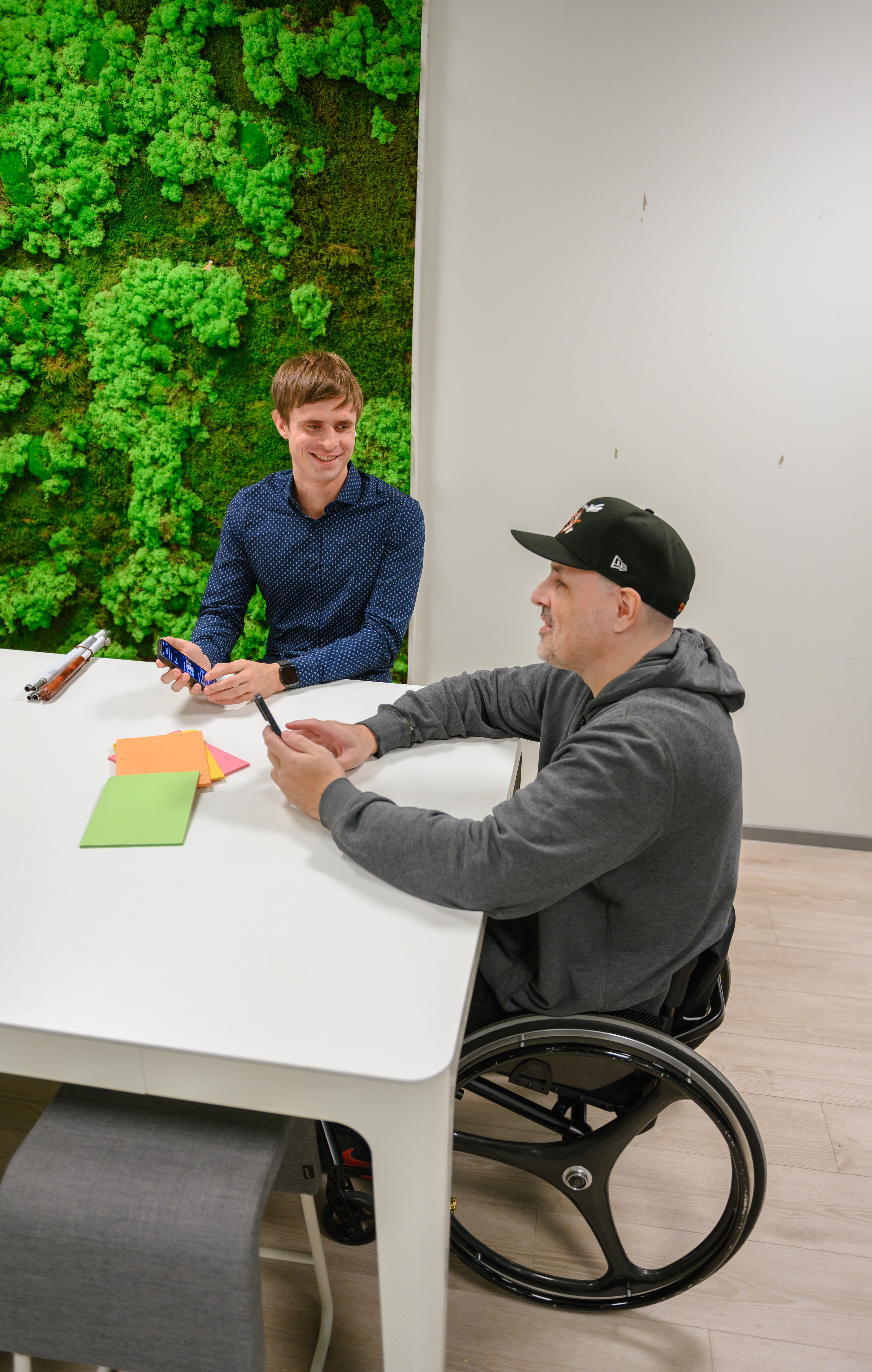 To menn sitter ved et møteromsbord. Mannen nærmest kamera er i rullestol og har caps på hodet. Begge smiler. De holder hver sin mobiltelefon i hånda. På veggen i bakgrunnen er det en veggdekorasjon som ser ut som en eng med blomster.