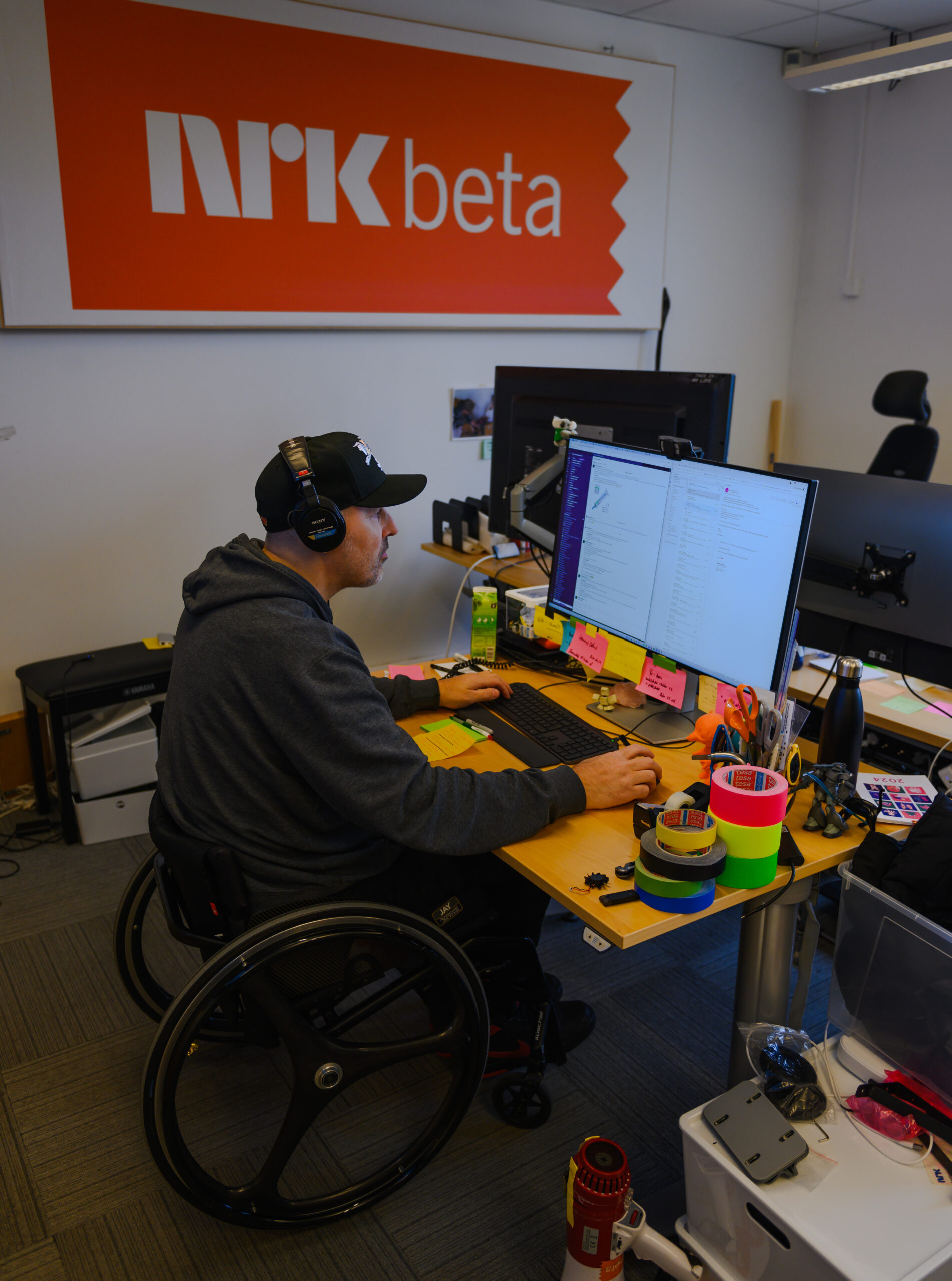 Bildet viser en mann i rullestol som sitter ved en pult og jobber ved en pc-skjerm. Han har flere store tape-ruller på pulten. Mannen har på seg hettegrenser og caps og han hører på noe via hodetelefoner. På veggen i bakrgunnen er det et stort skilt der det står: NRK beta. 