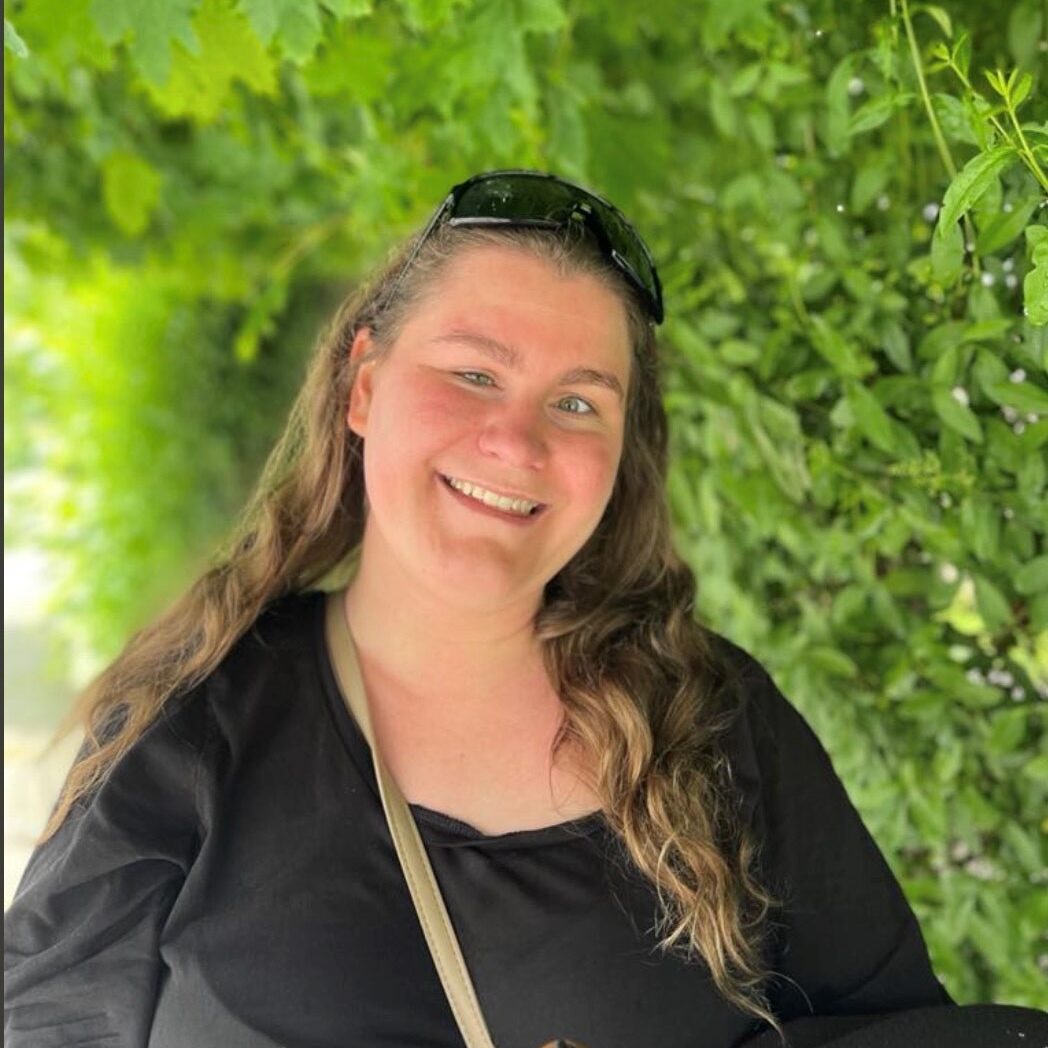 Bildet viser Ragna Kornelia Unstad Forså som smiler med åpen munn mot kamera. Hun har langt hår og solbriller på hodet. Hun har på seg en mørk topp. I bakgrunnen er det en hekk eller trør med grønne blader.